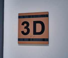 Close-up of an orange square sign with large black '3D' text and a dark horizontal stripe, mounted on a light gray wall.