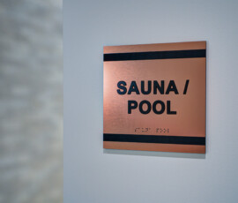 Copper plaque on gray wall reading 'SAUNA / POOL' with a black top stripe for contrast.