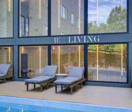 Outdoor lounge area beside a modern glass-walled building with blue-gray framing; blue cushioned lounge chairs and small side tables by a pool edge.