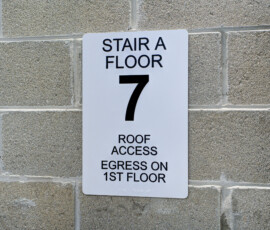 White rectangular sign on a gray brick wall reading 'STAIR A FLOOR 7' with smaller text about roof access and egress on the 1st floor, directing stairs.