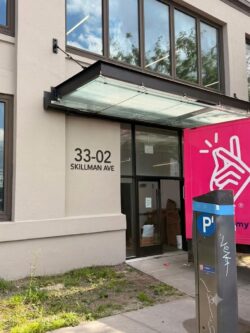 Entrance to a building at 33-02 Skillman Ave with a glass canopy; a pink delivery truck is parked nearby and a blue/black parking kiosk is in the foreground.